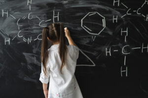 Cute little girl writing on a blackboard at a chemistry lesson in class. White coat and goggles.